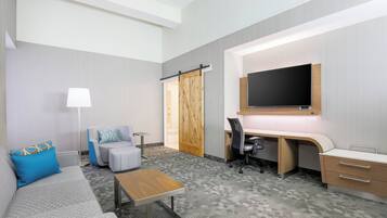 Quarto de hotel com sofá, escrivaninha, cadeira, televisão e porta de correr.