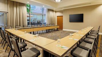 Un salón de conferencias con una larga mesa, sillas, un televisor y una ventana con cortinas.