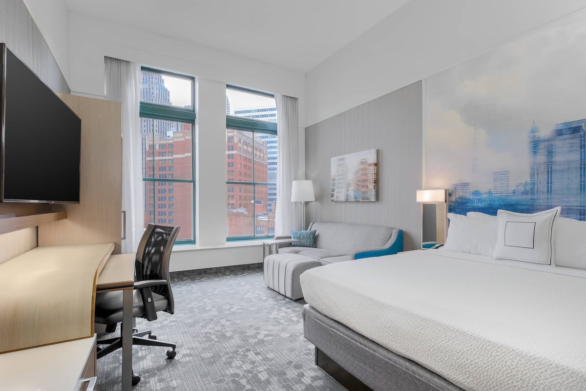 A hotel room with a large bed, a desk, a chair, a TV, and a window with a city view.