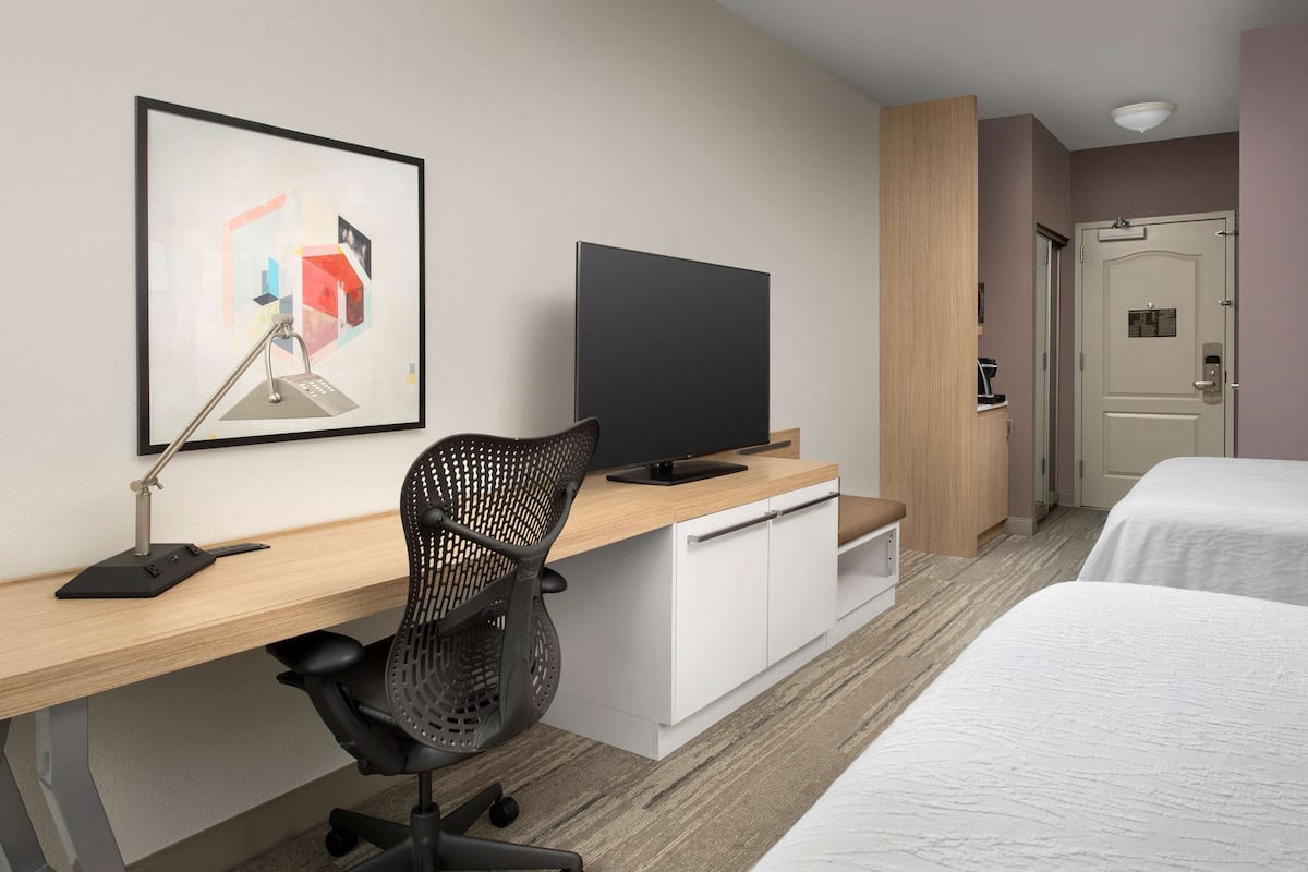 Une chambre d’hôtel avec un bureau en bois, un fauteuil de bureau noir, un téléviseur à écran plat, un lit avec une literie blanche et une œuvre d’art abstraite encadrée accrochée au mur.