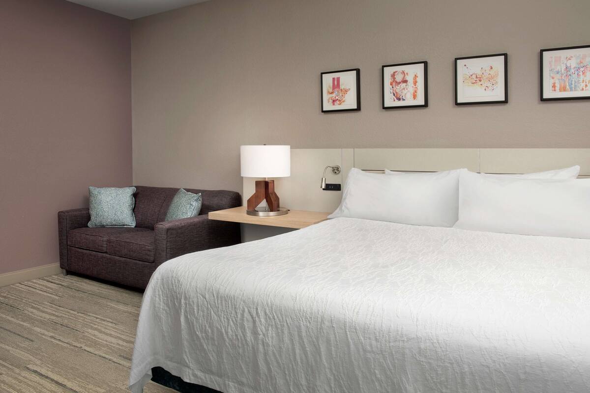 Une chambre d’hôtel avec un grand lit, un canapé et une table de chevet avec une lampe. Quatre tableaux encadrés ornent le mur.