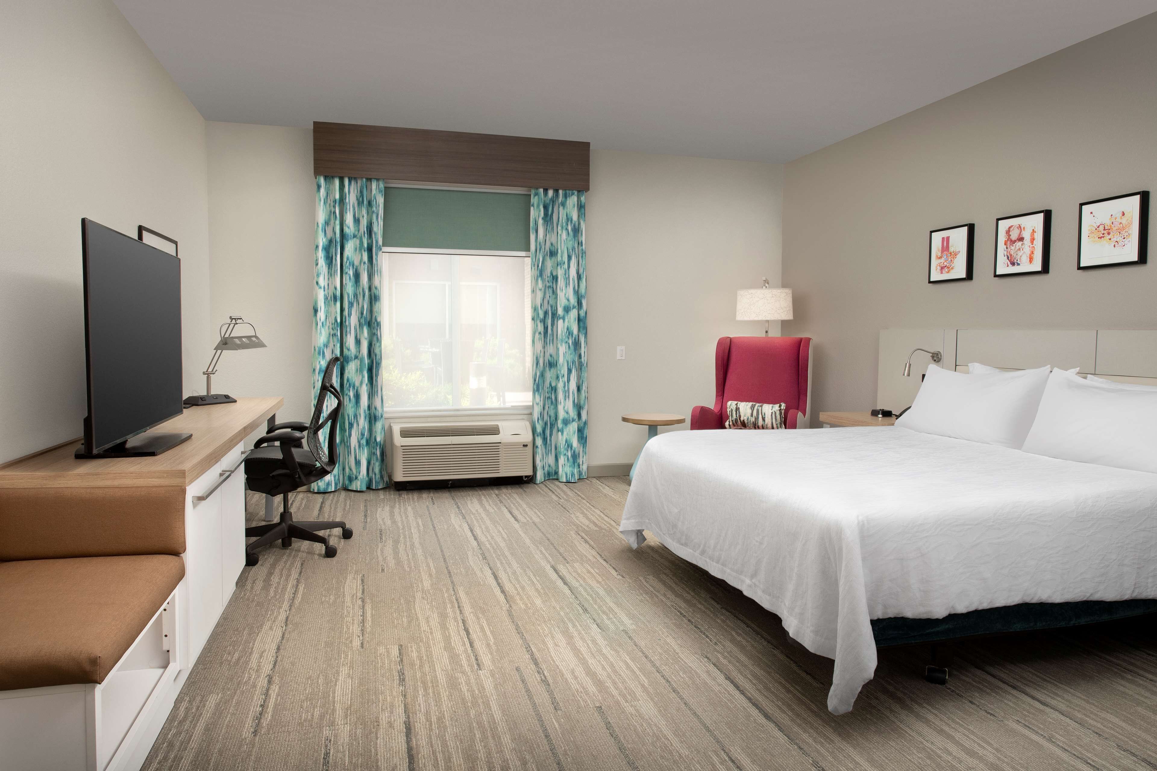 Habitación de hotel con cama, escritorio con computadora, silla, televisor y ventana con cortinas.