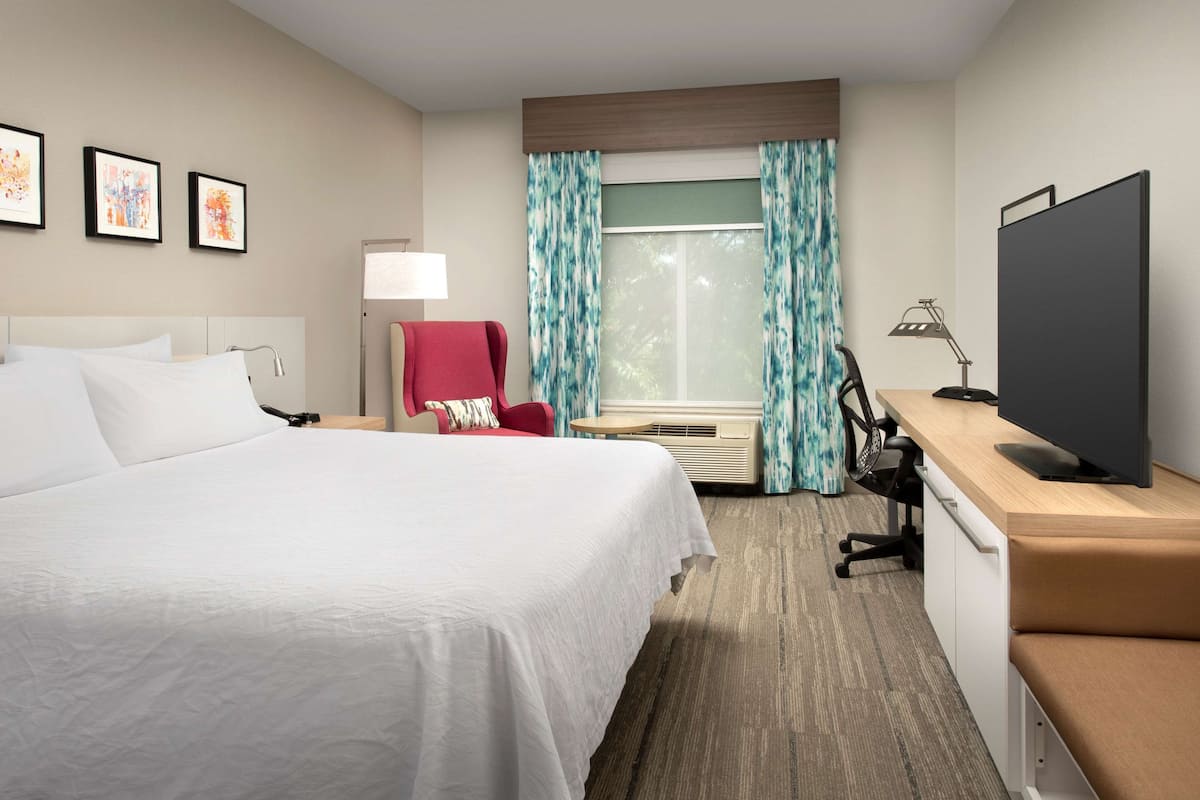 Une chambre d’hôtel avec un grand lit, un bureau avec un ordinateur, une télévision, un fauteuil rouge et une fenêtre avec des rideaux.