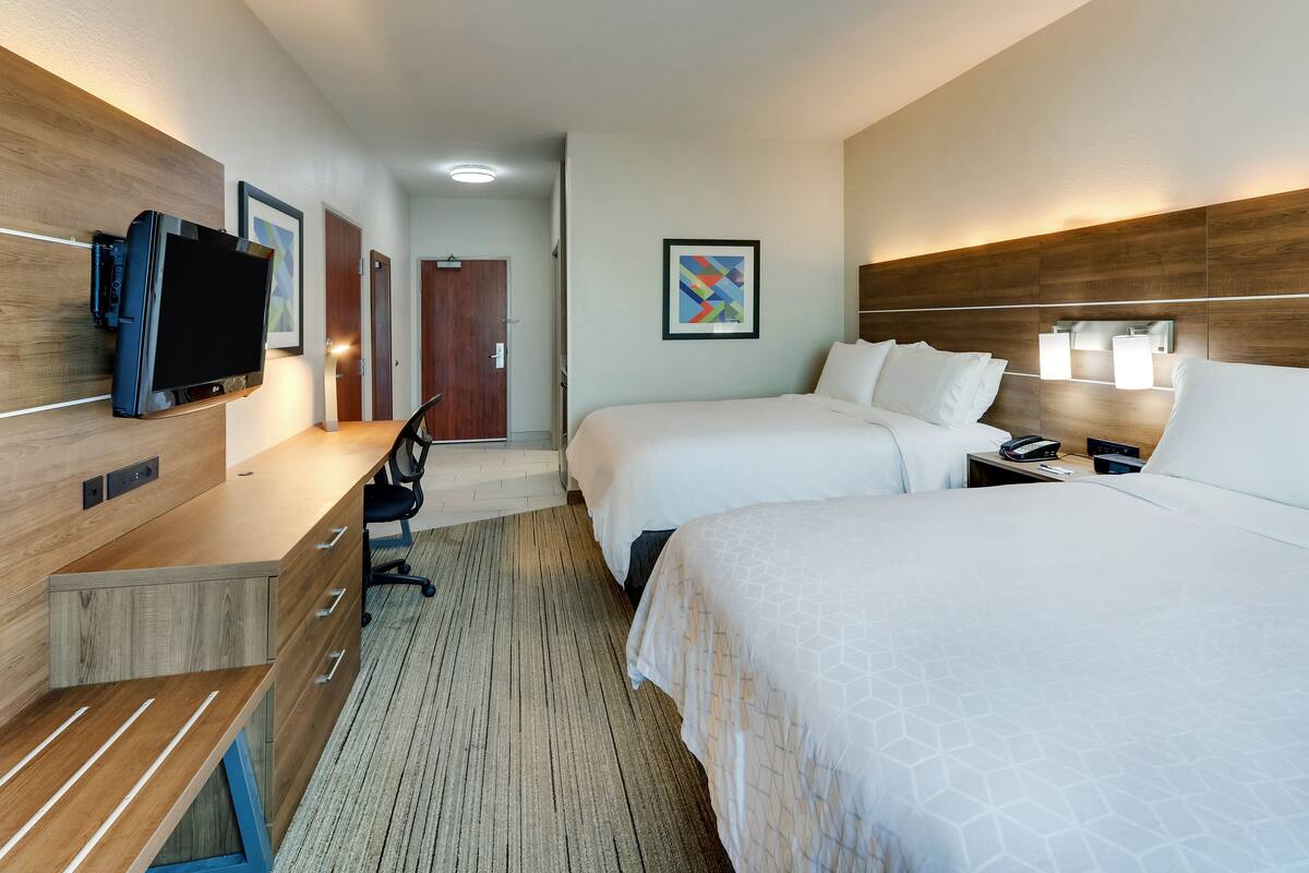 Habitación de hotel con dos camas, un escritorio, un televisor de pantalla plana, una silla y una lámpara de pared.