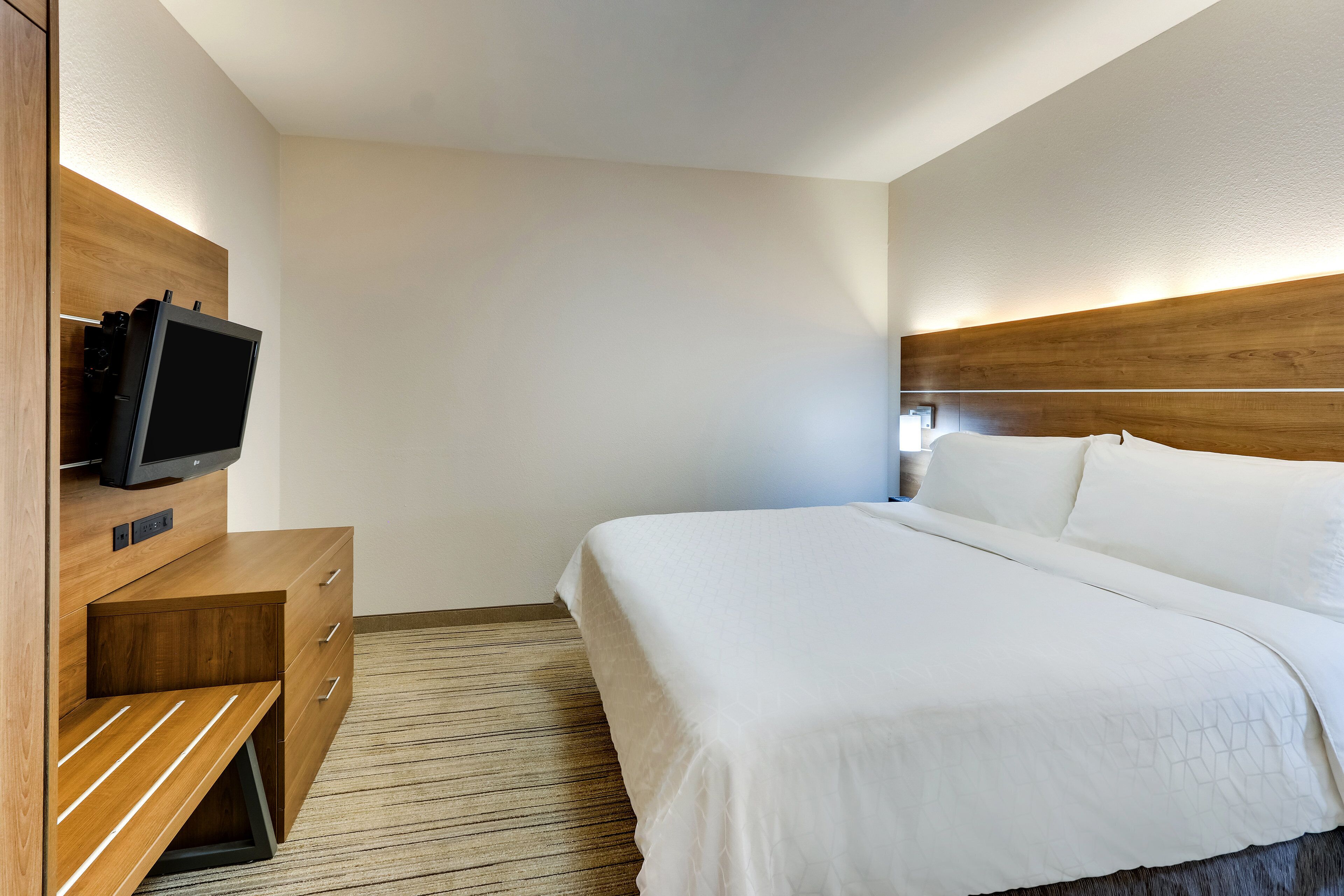 A hotel room with a bed, a wooden nightstand, a television, and a wall-mounted light.