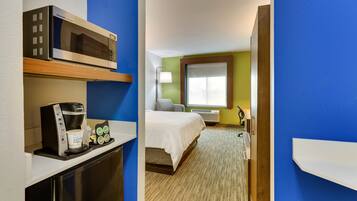 Quarto de hotel com cozinha compacta, cama com edredom branco, janela com persianas e uma pequena mesa de cabeceira.