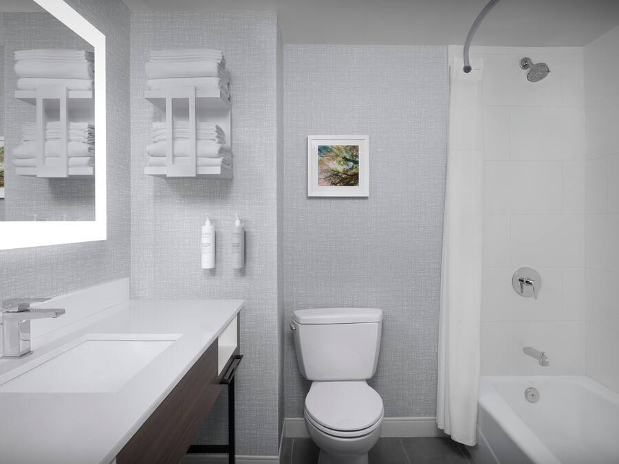 Une salle de bain moderne avec un lavabo, des toilettes et une baignoire, tous de couleur blanche. Des serviettes sont suspendues sur un porte-serviettes et un tableau encadré est accroché au mur.