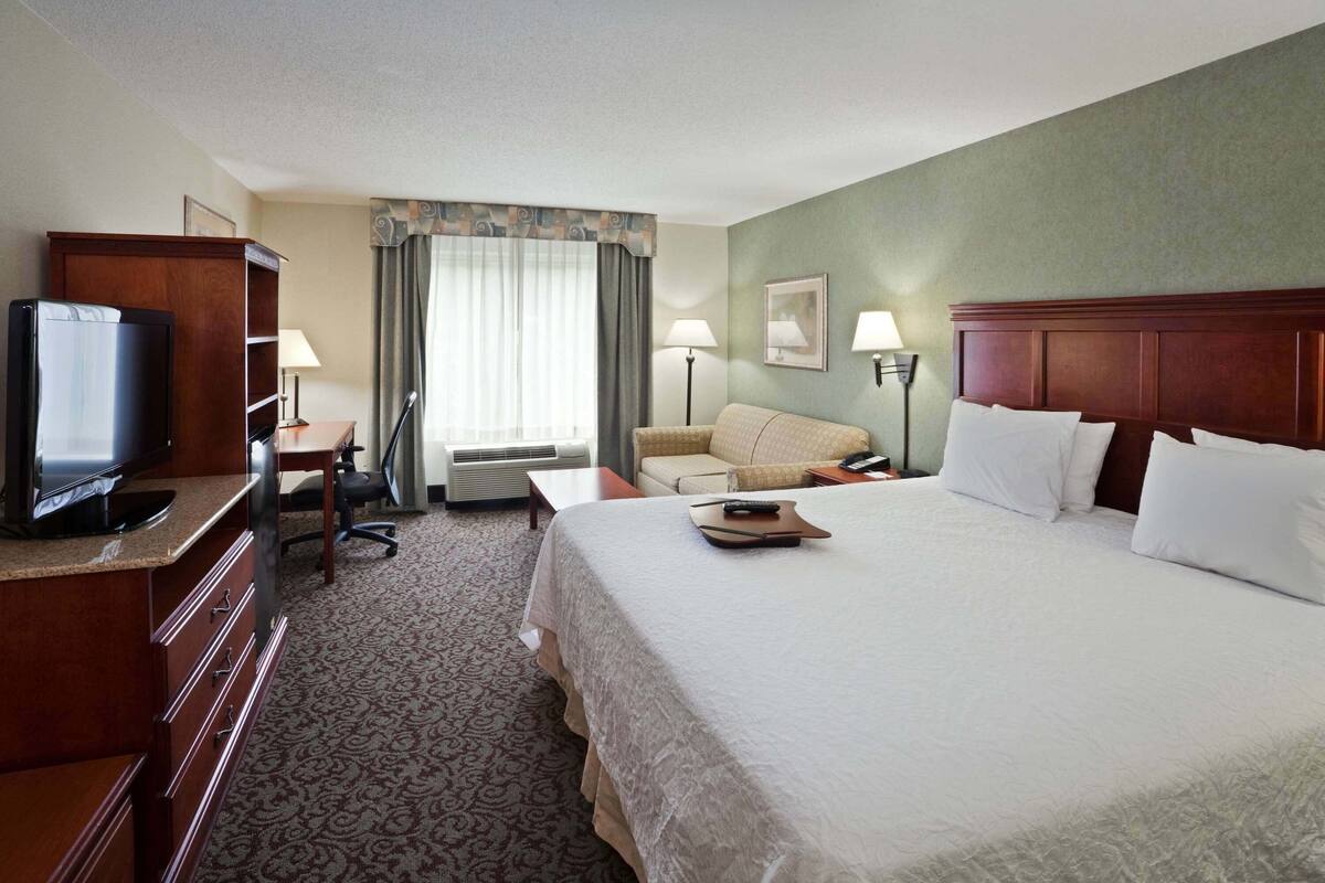A hotel room with a bed, a desk with a television, a chair, a couch, and two lamps.