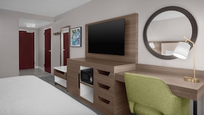 A modern hotel room with a large bed, a wooden desk with a flat-screen TV, a green chair, a round mirror, and a small shelf with a microwave.