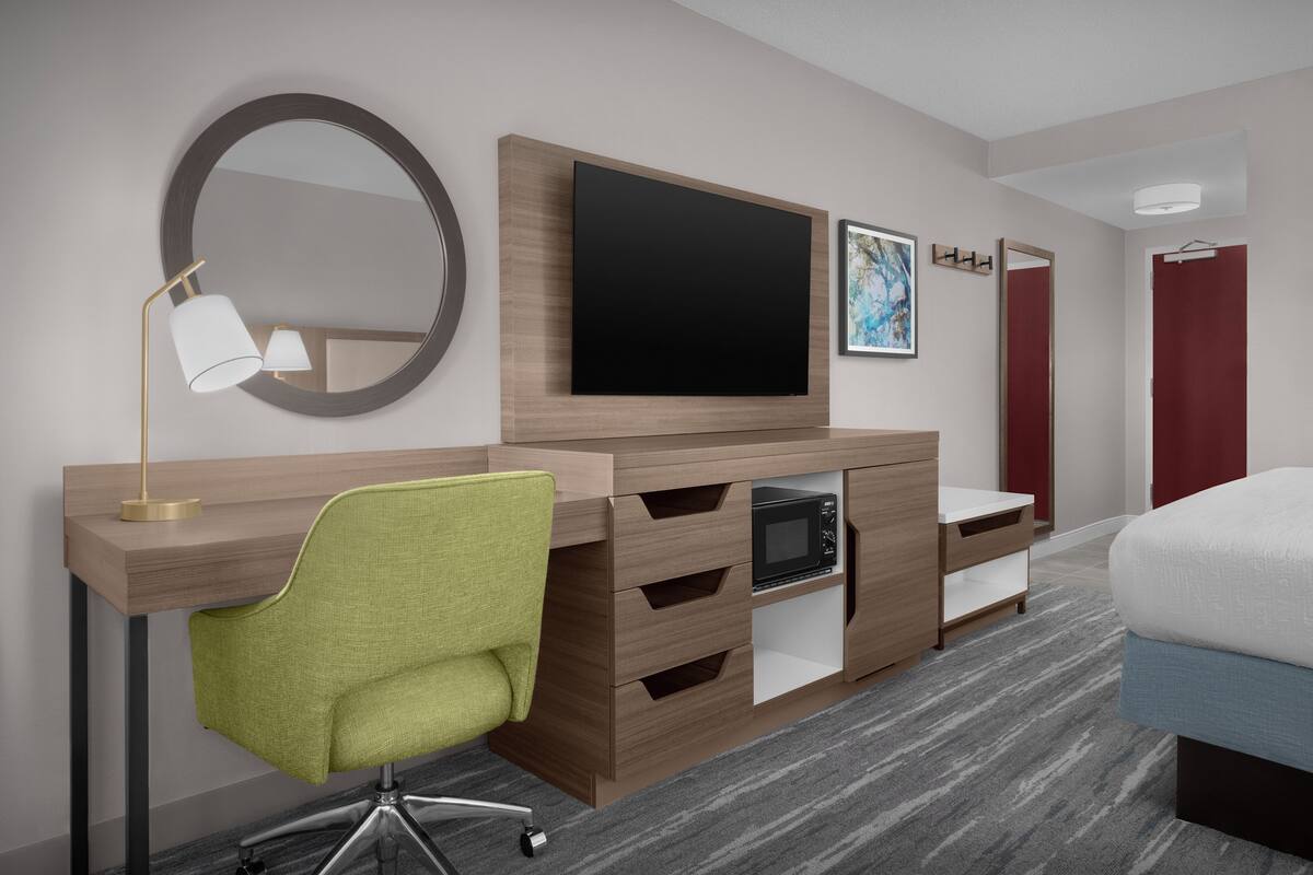 A modern hotel room with a wooden desk, a green chair, a flat-screen TV, a microwave, a small cabinet, and a bed.