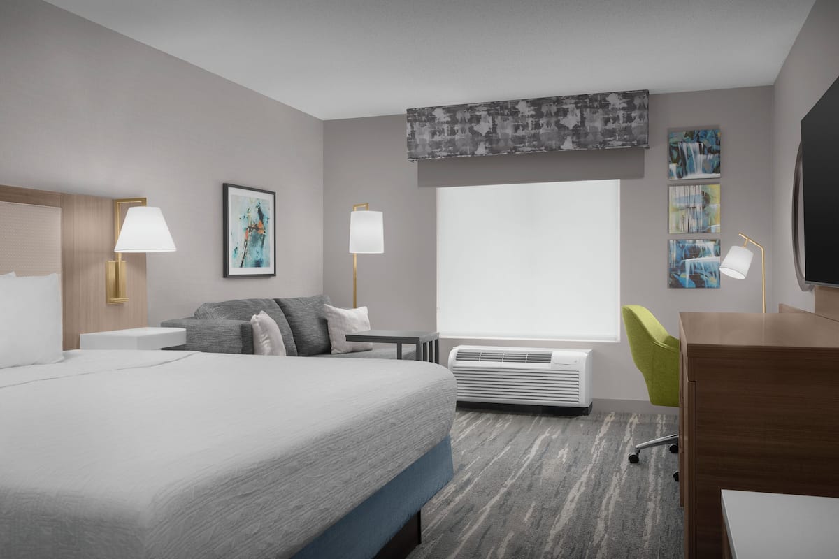 A modern hotel room with a large bed, a desk, a chair, a window, and a wall-mounted TV.