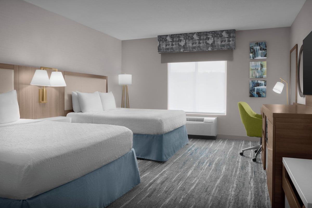 A hotel room with two beds, a desk, a chair, a TV, and artwork on the wall.