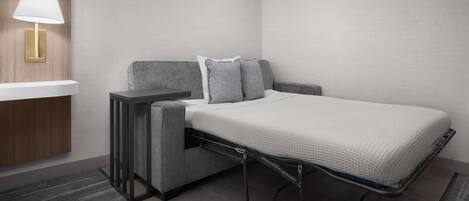 A compact hotel room with a bed, a bedside table, a lamp, and a small seating area.