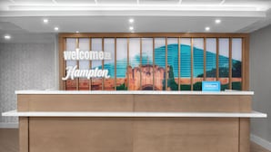 A reception desk with a "Welcome to Hampton" sign, a Hilton Honors logo, and a mural of a building.