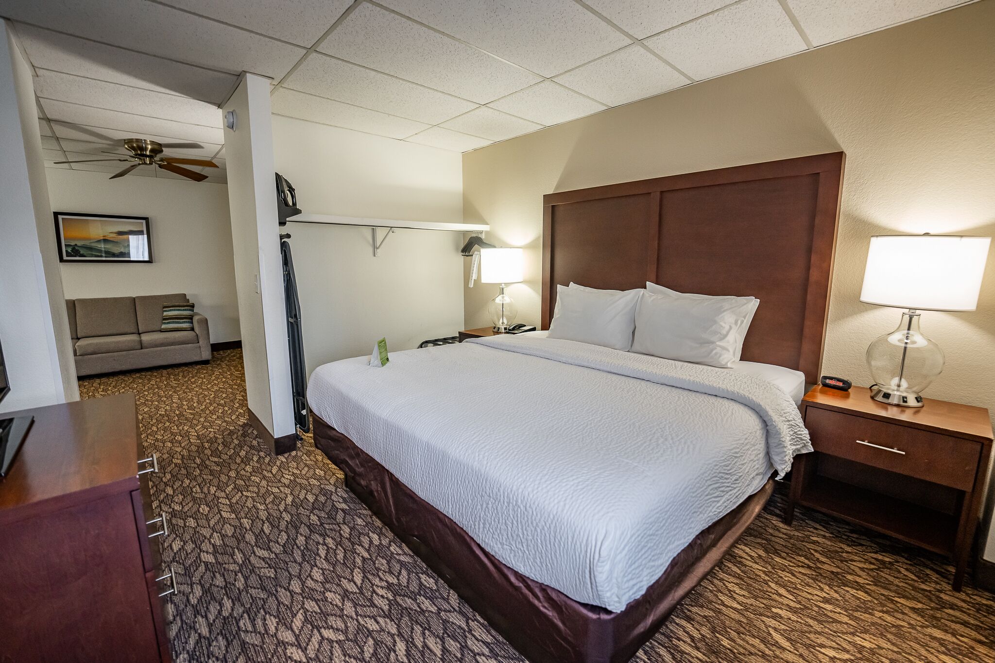 A hotel room with a large bed, a wooden headboard, two bedside lamps, a nightstand, a desk, a ceiling fan, and a couch.
