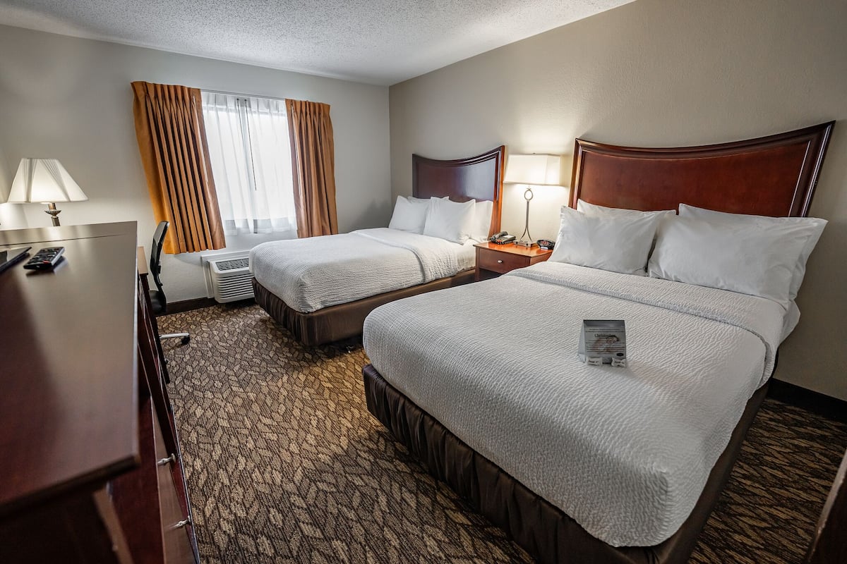 Quarto de hotel com duas camas, uma escrivaninha, uma cadeira, um abajur e uma janela com cortinas.