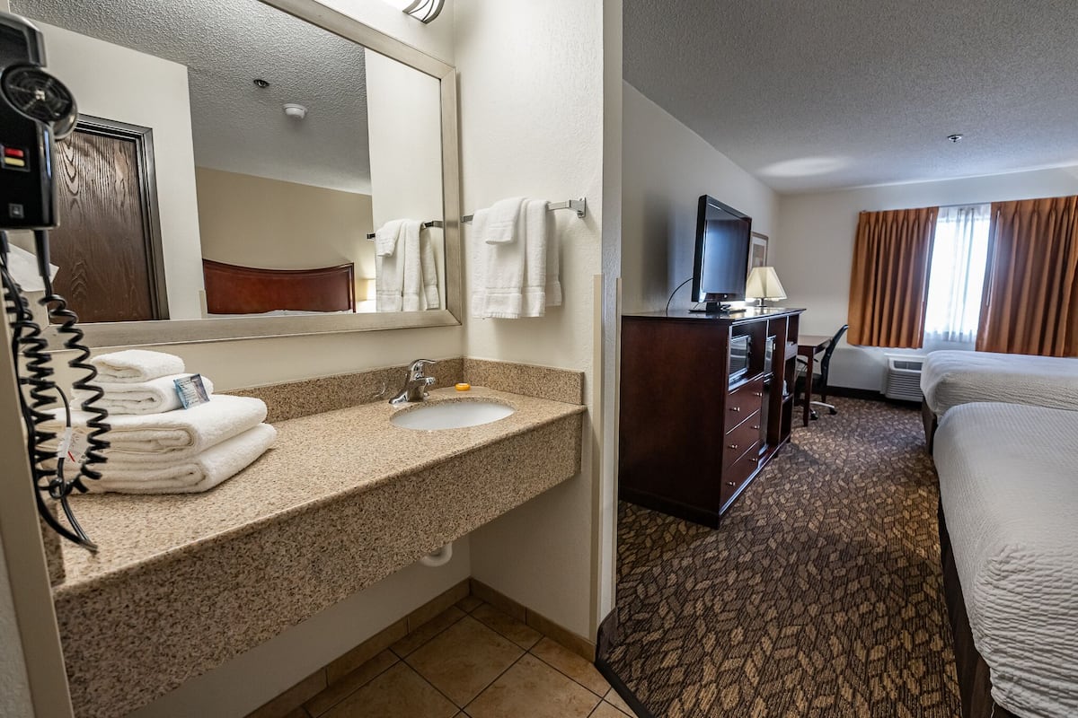 Quarto de hotel com pia de bancada de granito, espelho, telefone, escrivaninha com televisão e cama com carpete estampado.