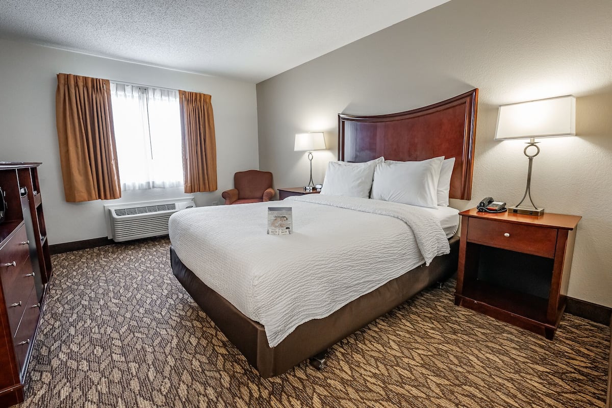 Quarto de hotel com uma cama grande, duas luminárias de cabeceira, uma mesinha de cabeceira de madeira, uma cadeira e uma escrivaninha com abajur.