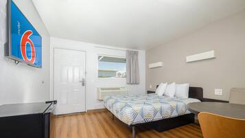 A hotel room with a bed, a desk, a chair, a window with curtains, and a wall-mounted air conditioner.