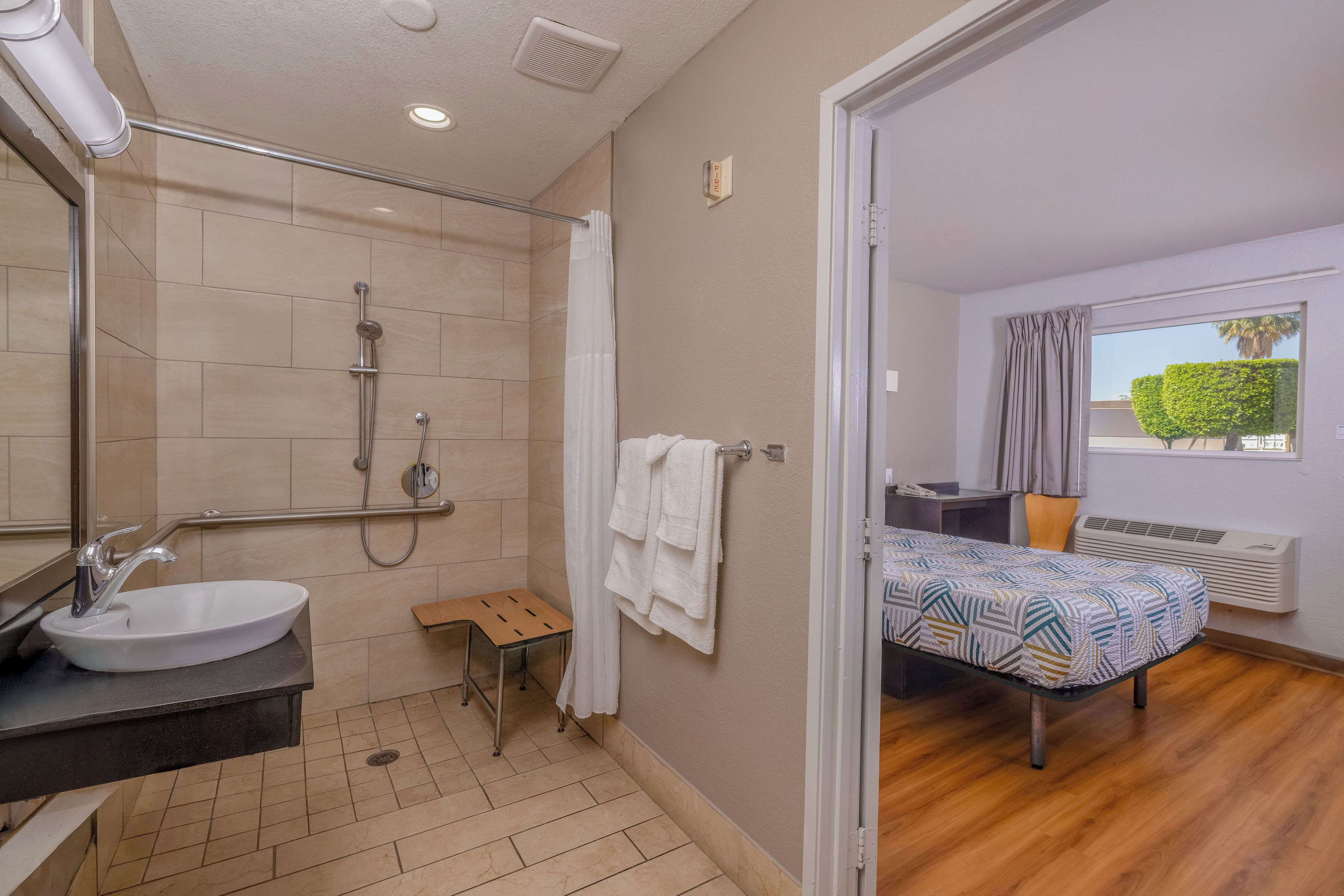 A bathroom with a sink, shower, and a small stool. There is a towel rack with towels and a window with curtains.