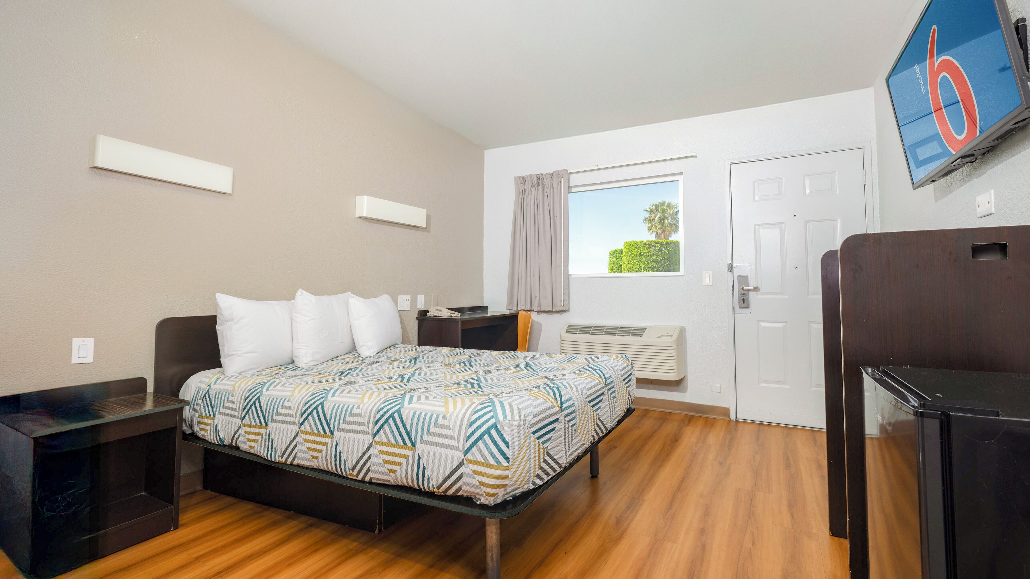 A hotel room with a bed, bedside tables, a desk, a TV, and a window with a view of greenery.