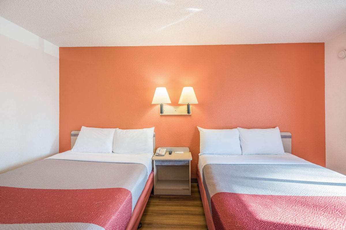 A hotel room with two beds, a nightstand with a lamp, and a wooden floor.