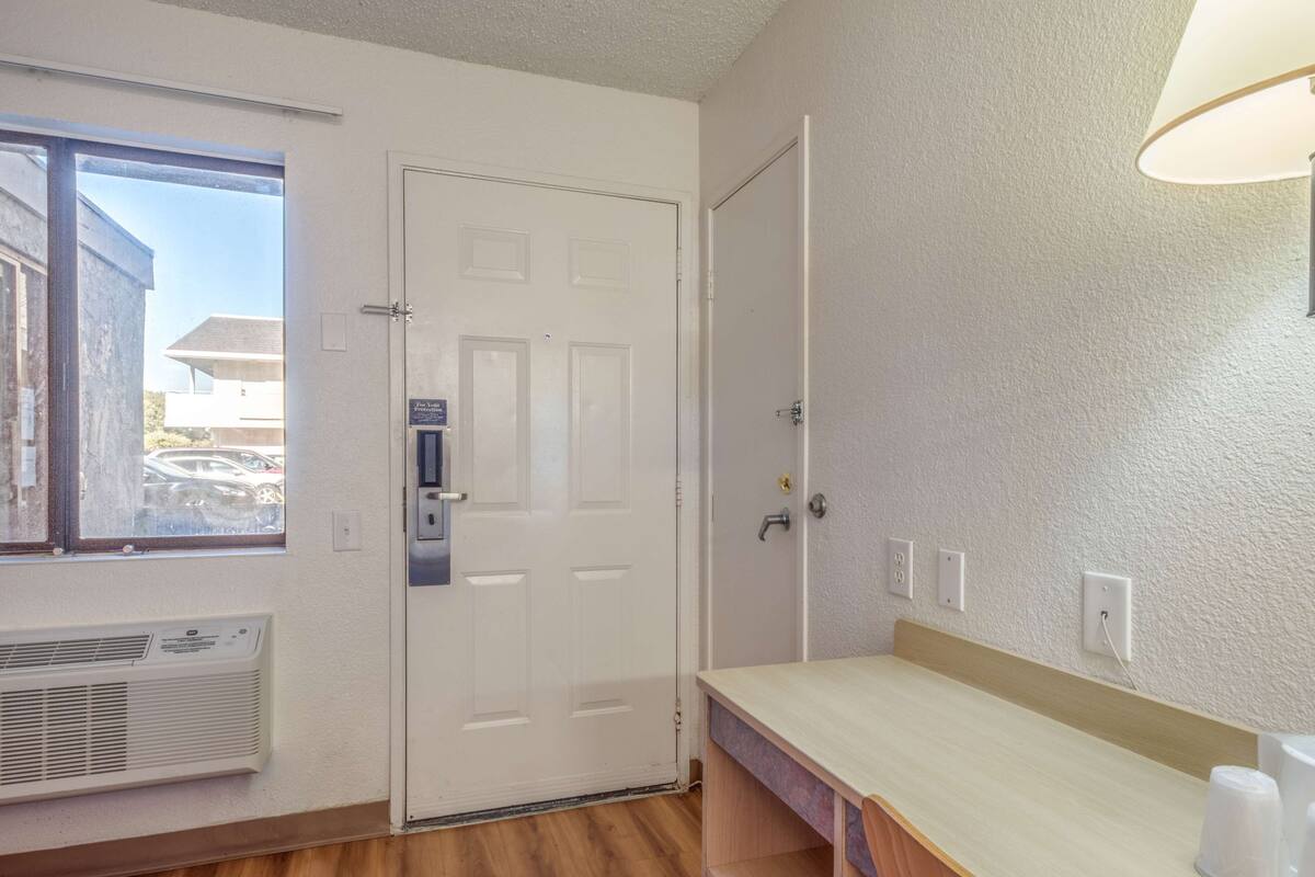 A room with a white door, a window with a view of parked cars, a wooden bench, and a wall-mounted lamp.