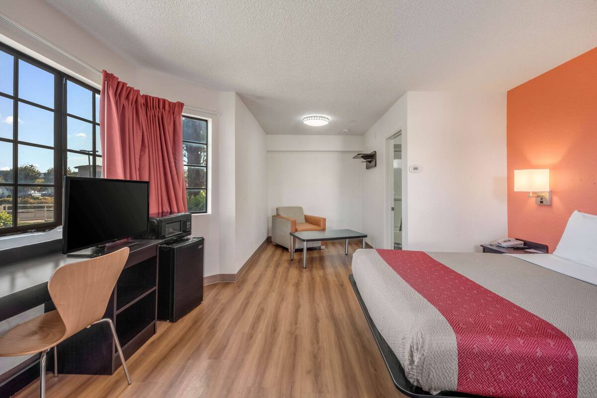 A hotel room with a bed, a desk with a TV, a chair, a small table, and a window with curtains.