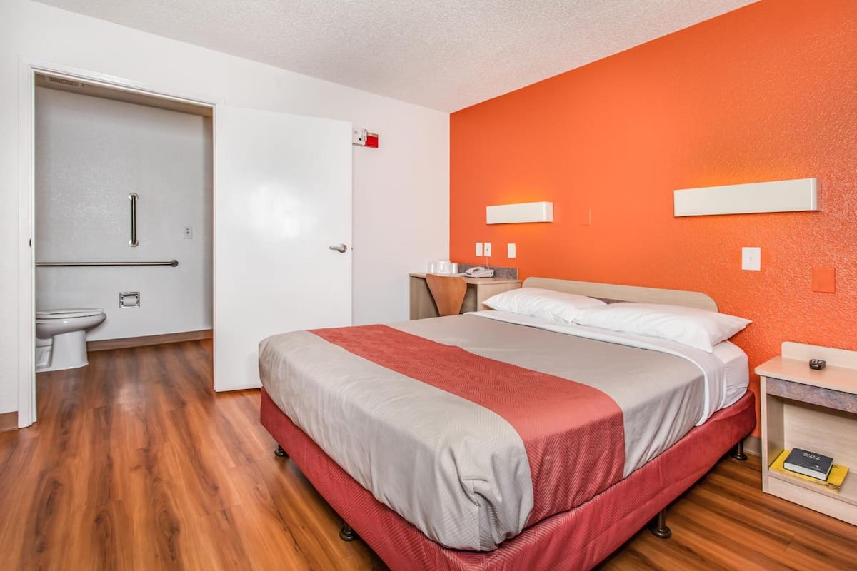 A hotel room with a bed, bedside tables, a nightstand, a red wall, and a door leading to a bathroom.