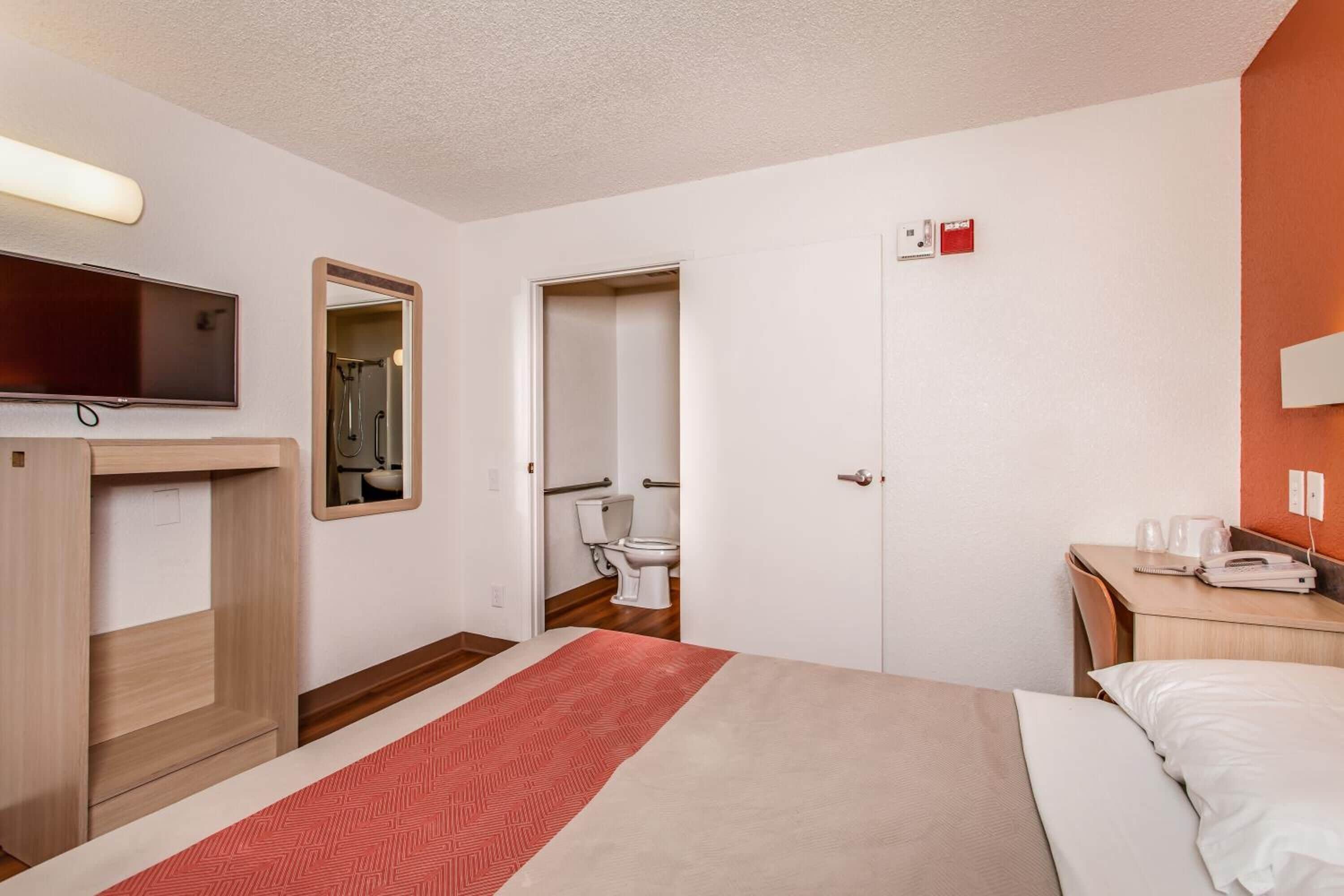 A hotel room with a bed, a desk with a telephone, a mirror, and a television mounted on the wall.