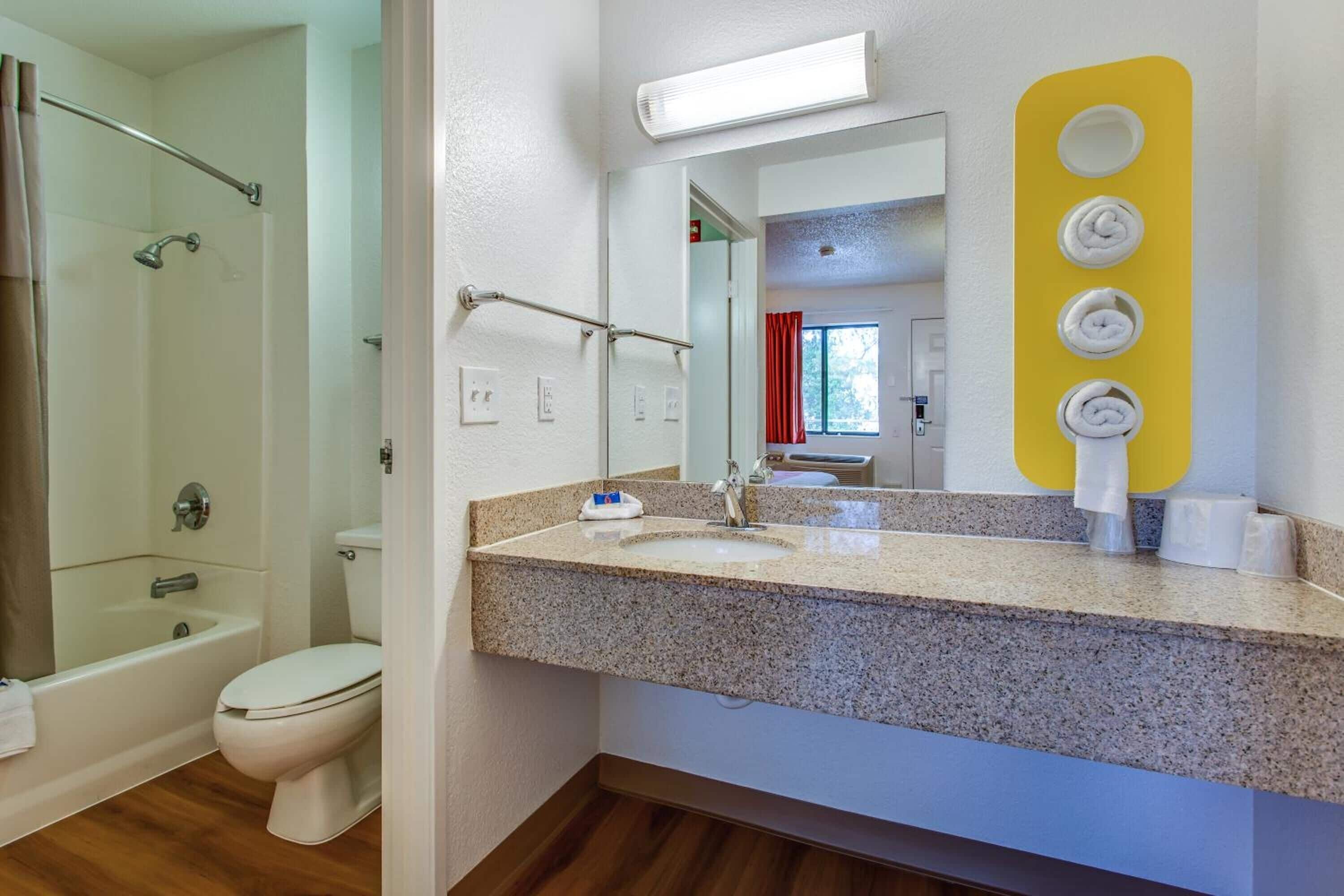 A bathroom with a shower, toilet, granite countertop, sink, and a mirror with a yellow towel holder.