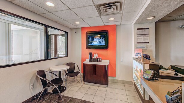 A reception area with a TV mounted on the wall, a small table with chairs, a cabinet with a coffee machine, and a counter with a phone and computer. - Motel 6 Niantic, CT - New London (Niantic)
