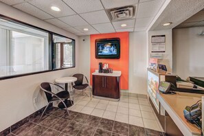 A reception area with a TV mounted on the wall, a small table with chairs, a cabinet with a coffee machine, and a counter with a phone and computer. - Motel 6 Niantic, CT - New London (Niantic)
