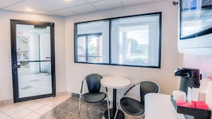 A small, modern room with a round table and two chairs, a window with a view, and a trash bin with a coffee machine. - Motel 6 Niantic, CT - New London (Niantic)
