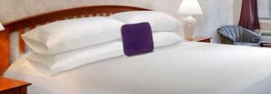 A neatly made bed with white linens and a purple pillow, a wooden headboard, a bedside lamp, and a chair with a round table beside the bed.