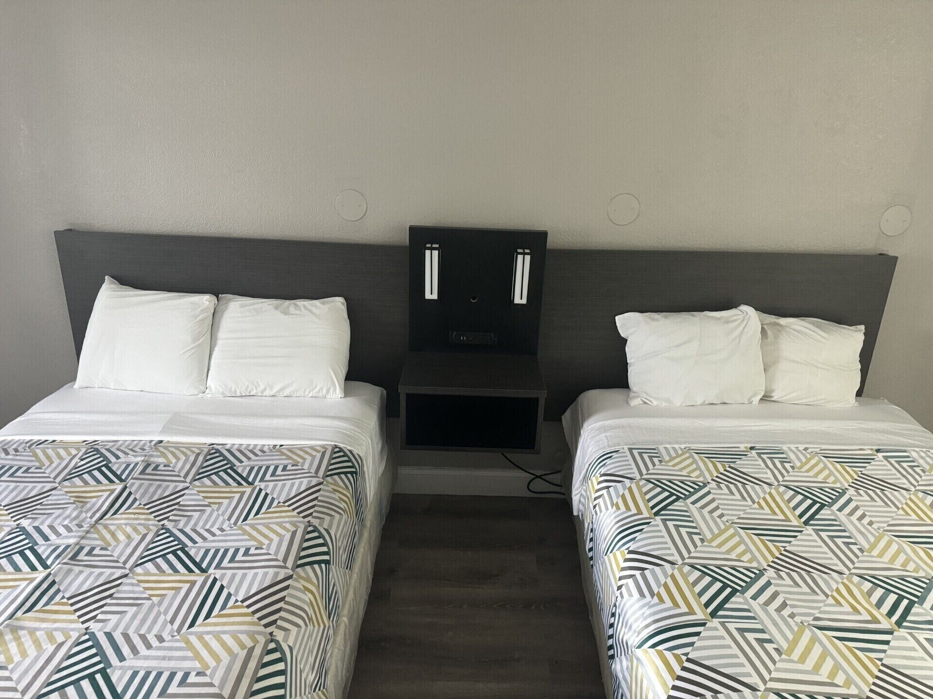 Habitación de hotel con dos camas, cada una con una colcha estampada y almohadas blancas. Entre las camas hay una mesita de noche con una lámpara y una pequeña mesa.