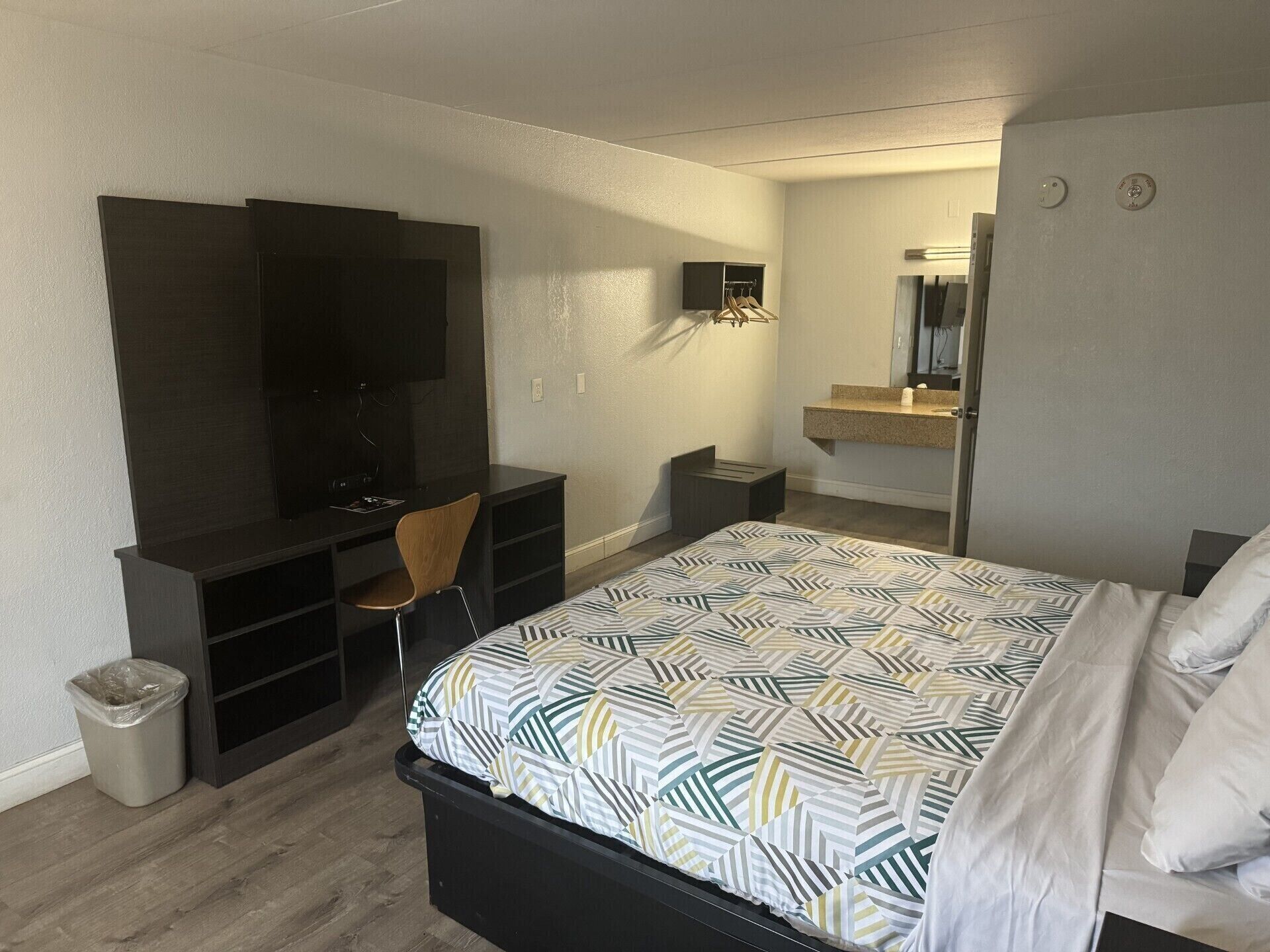 Un dormitorio con cama, escritorio, silla, televisión, espejo y un cesto de basura pequeño.