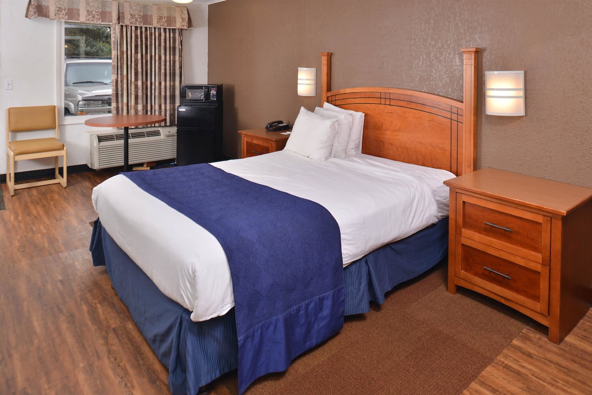 Ein Hotelzimmer mit einem großen Bett, einem hölzernen Nachttisch, einem Schreibtisch mit Stuhl, einem Heizgerät und Blick auf ein parkendes Auto durch das Fenster.