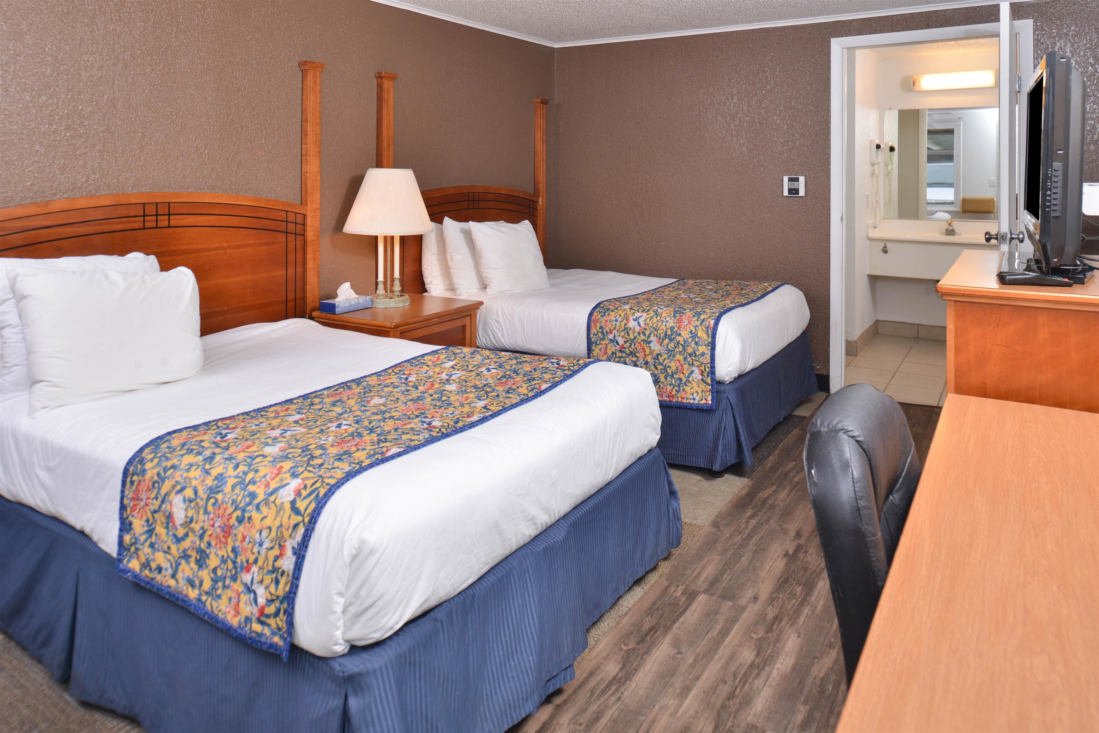 A hotel room with two beds, a desk, a chair, a lamp, and a television.