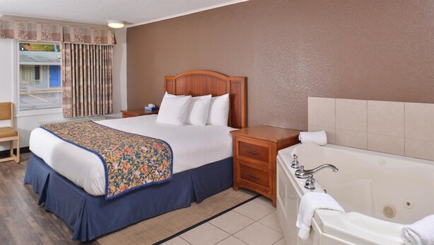 A hotel room with a bed, a nightstand, a dresser, a bathroom with a bathtub, and a window with curtains. - Americas Best Value Inn & Suites Jackson, MI (Jackson)