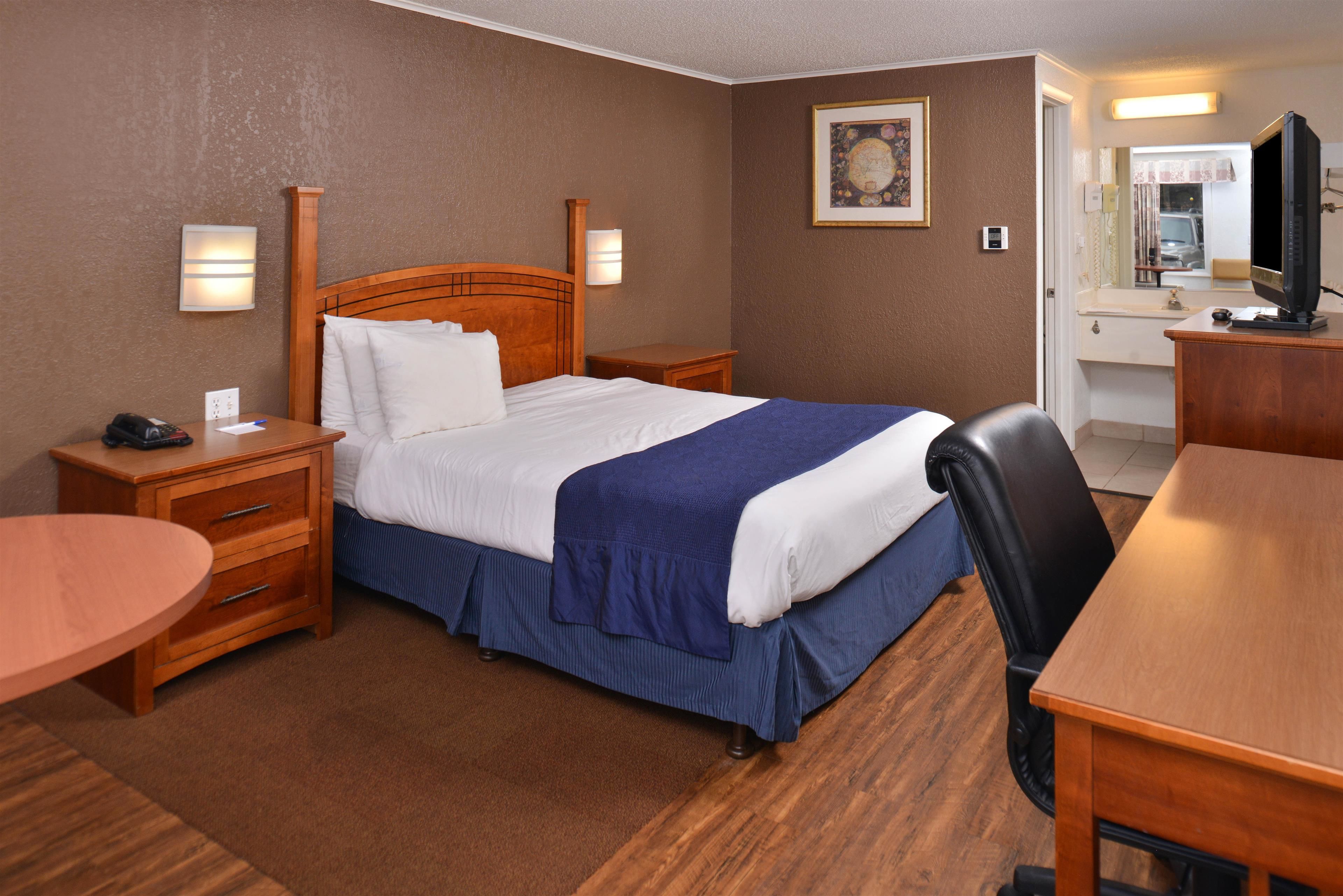 A hotel room with a wooden bed, a desk, a chair, a small table, and a television.