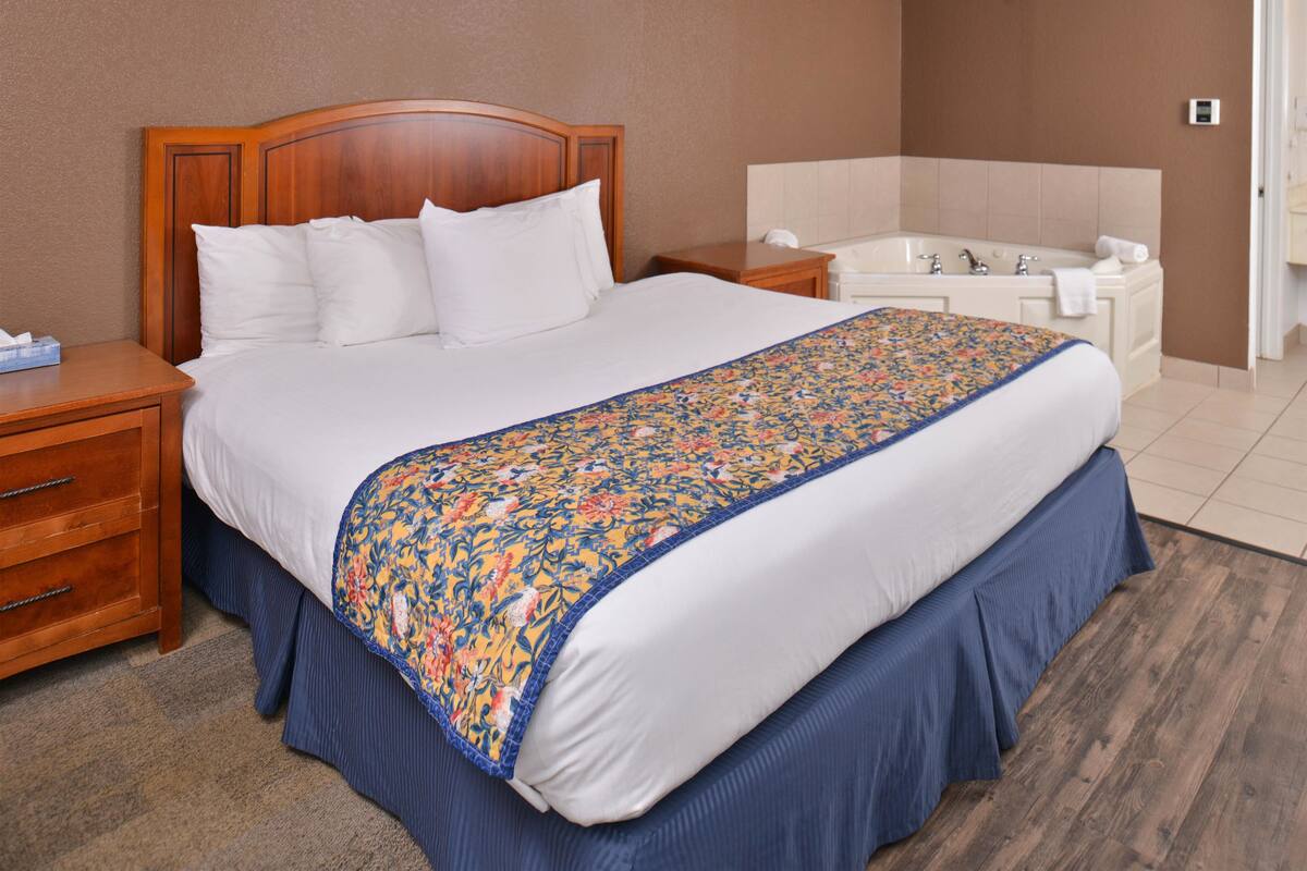 Ein Hotelzimmer mit einem großen Bett, einem Holz-Kopfteil, einem blumigen Bettüberwurf, einem Nachttisch und einem Bad mit Badewanne und Waschbecken.