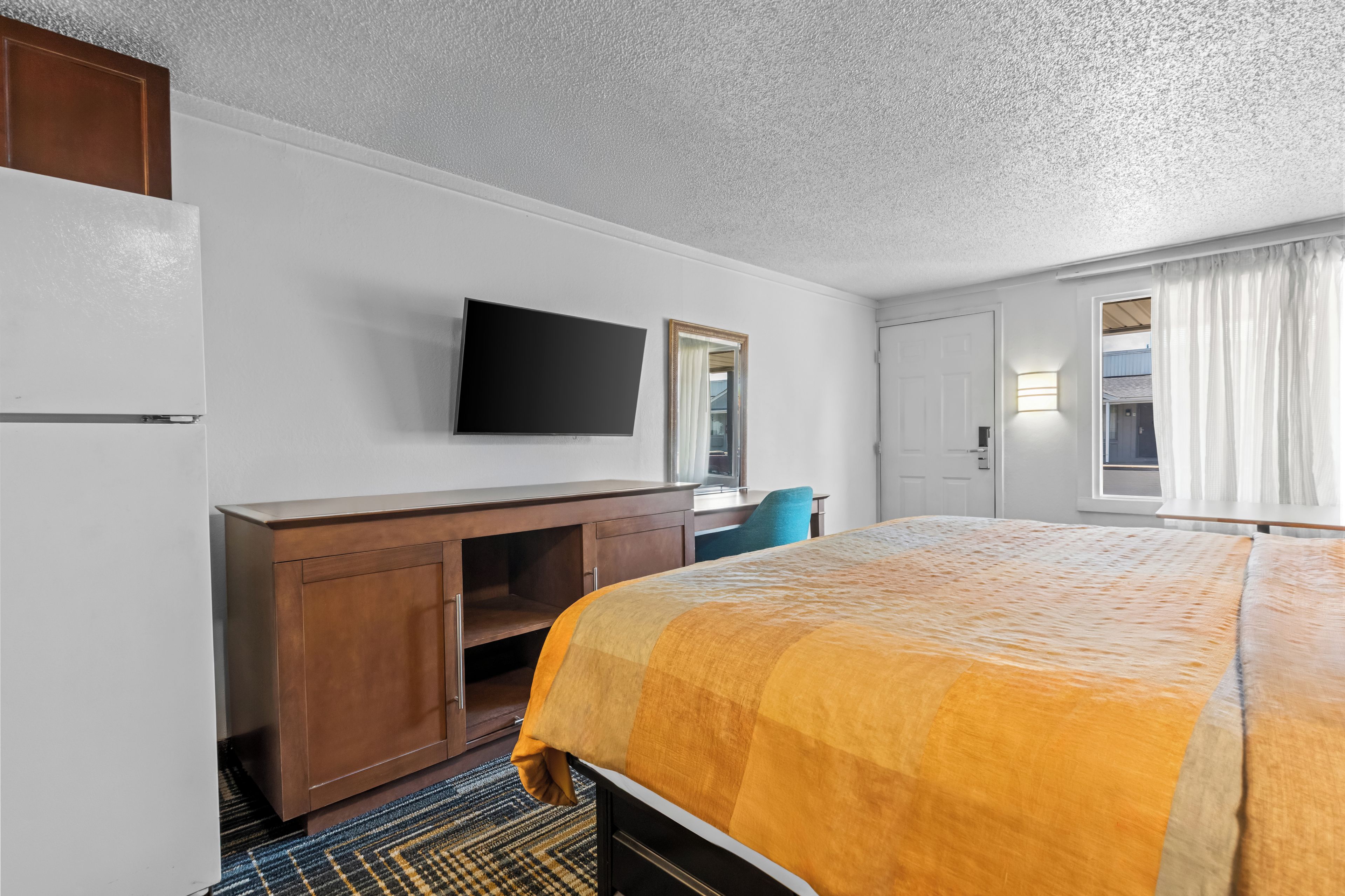 A hotel room with a bed, a flat-screen TV mounted on the wall, a wooden desk, a chair, and a window with curtains.