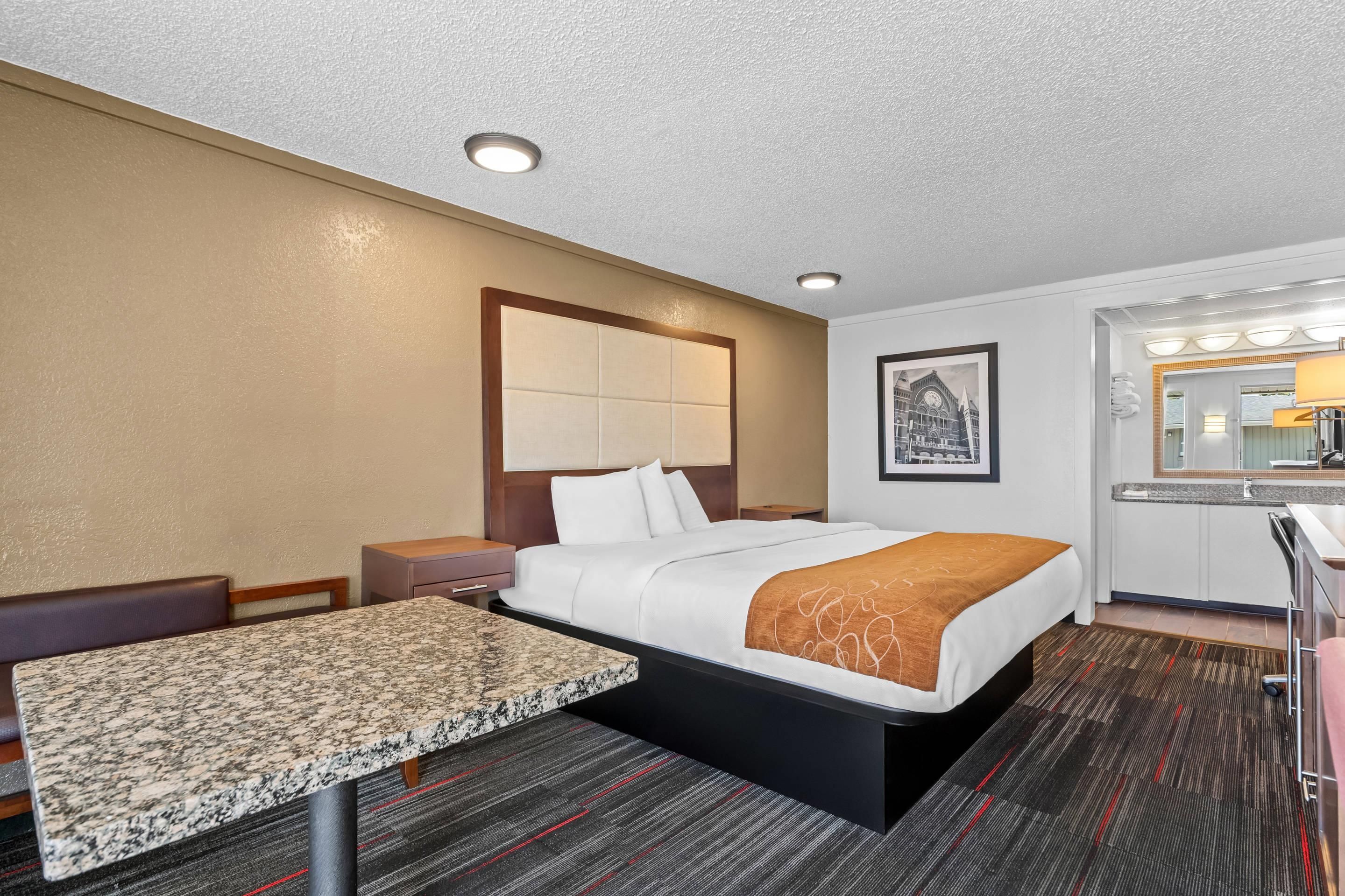 A hotel room with a bed, a desk, a chair, a nightstand, a mirror, and a framed picture on the wall.