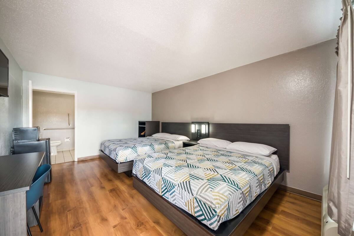 A modern hotel room with two beds, a desk, and a small kitchenette.