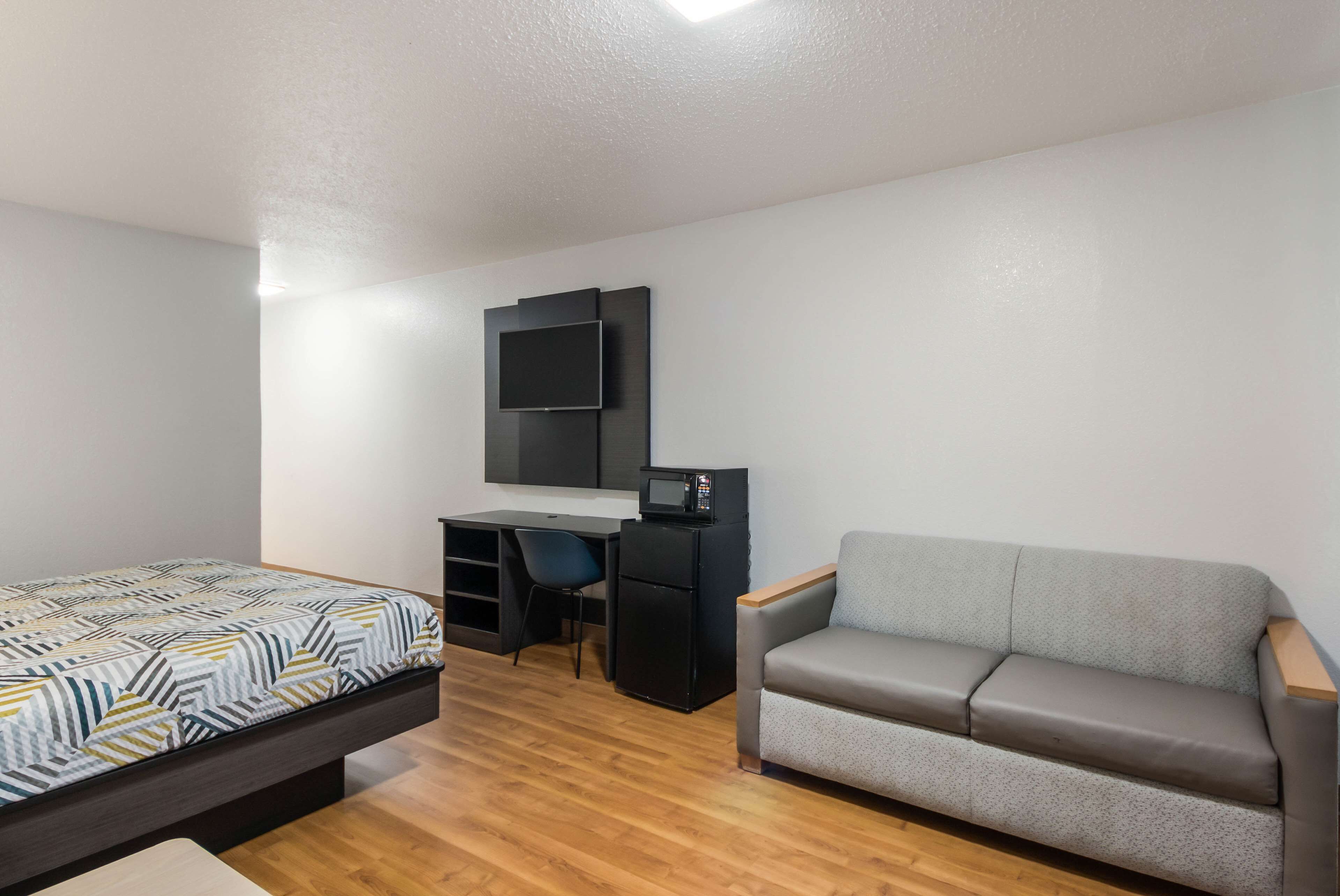 A compact room with a bed, a desk with a chair, a sofa, and a TV mounted on the wall.