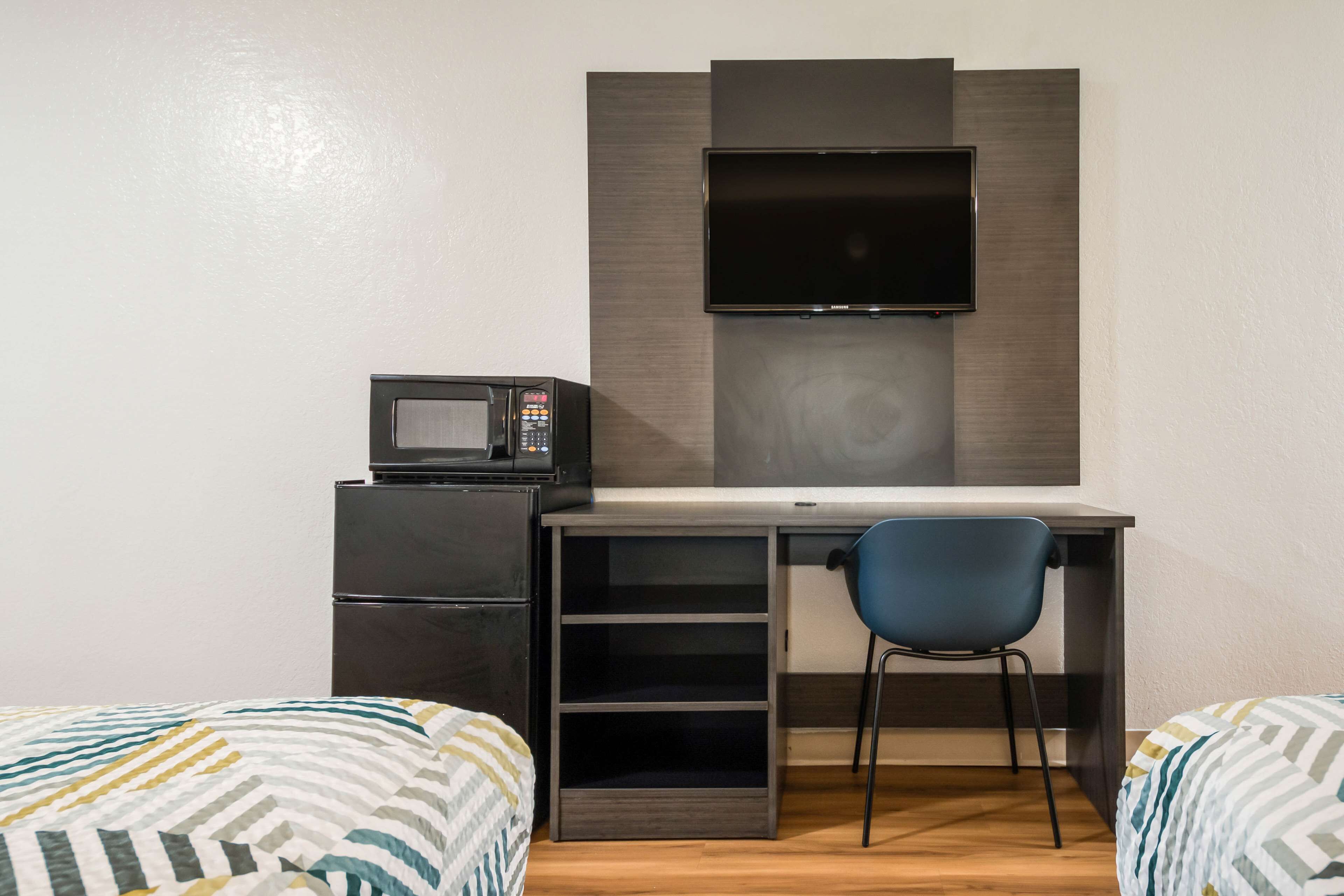 A compact room with a desk, a flat-screen TV mounted above it, a microwave, and a chair. There are two beds with patterned bedding.