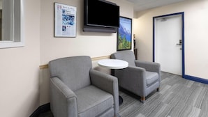 A room with two grey armchairs, a small round table, a mounted flat-screen TV, and a door with a blue frame. - Travelodge by Wyndham Madison Heights MI (Madison Heights)