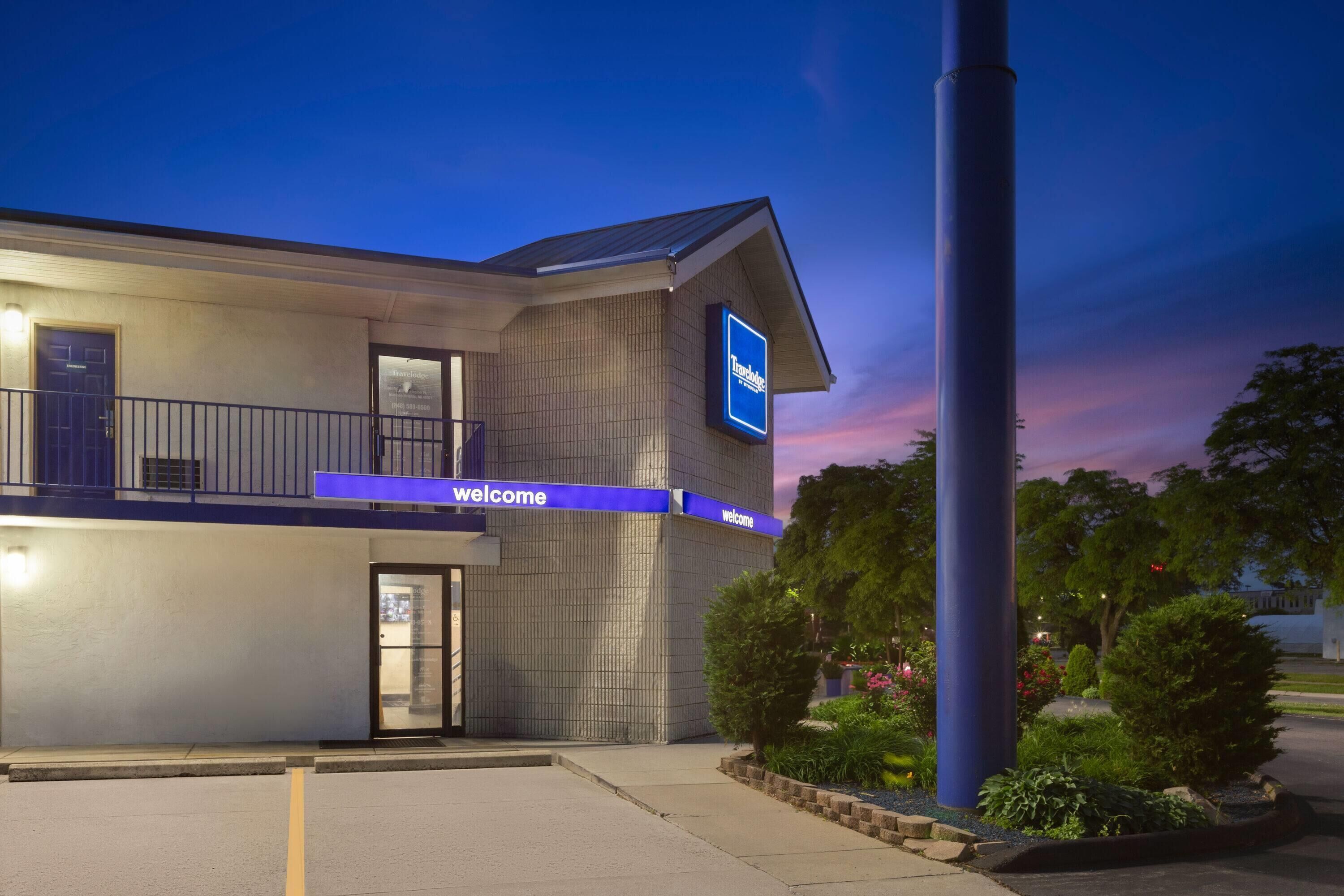 A modern hotel with a 'Welcome' sign, a balcony, and a well-maintained garden area.