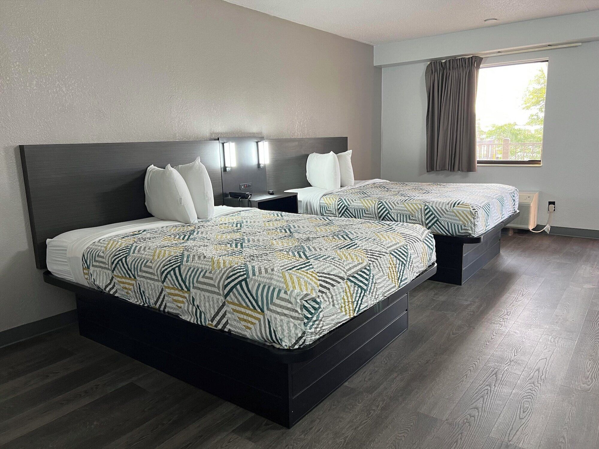 A hotel room with two beds, a large window with curtains, a bedside table with a lamp, and a wall-mounted air conditioning unit.